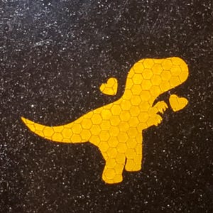 May include: A yellow dinosaur silhouette with a honeycomb pattern, holding two hearts in its front paws. The dinosaur is on a black background with a sparkly texture.