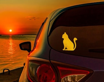 Könnte beinhalten: Eine goldene Silhouette einer Katze, die auf der Heckscheibe eines Autos sitzt. Das Auto ist an einem Gewässer mit einem Sonnenuntergang im Hintergrund geparkt.