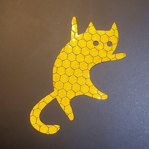 May include: A yellow cat-shaped sticker with a honeycomb pattern. The sticker has two black eyes and is made of reflective material.