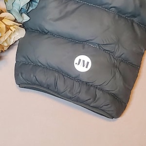 Puede incluir: Primer plano de la parte inferior de una chaqueta acolchada gris oscuro con un parche circular blanco que dice "JM" en letras negras.