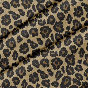 May include: A close-up of a tan fabric with a repeating leopard print pattern. The pattern features dark brown and black spots of varying sizes and shapes. The fabric is folded, showing the texture and drape of the material.