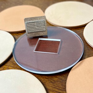 May include: A metal stamp with an American flag design sits on a clear acrylic circle, with a matching impression below. Several tan leather coasters surround the stamp and acrylic piece. The stamp is likely used for leathercraft or similar projects.