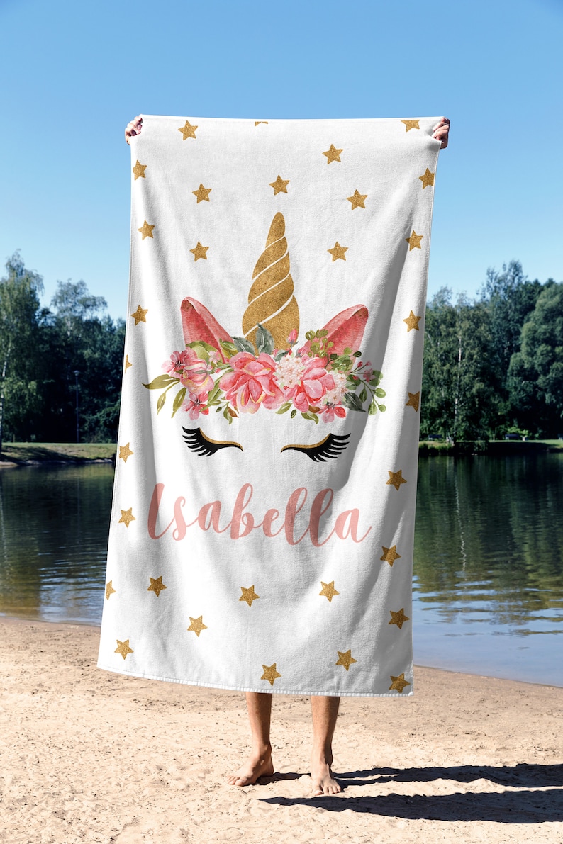 May include: White beach towel featuring a unicorn design with a gold horn, pink ears, and a floral crown. The towel has gold stars and the name "Isabella" in pink. The towel is held up on a beach.