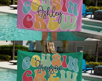 May include: Two beach towels with a retro design. The towels are green and pink with the words "Cousin Crew" in colorful, rounded letters. The name "Ashley" is written in black on the pink towel and the name "Greyson" is written in black on the green towel.