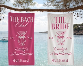 Puede incluir: Dos toallas de playa colgadas de una rama, una rosa y otra blanca. La toalla rosa dice "THE BACH Club" y "Emily's Bachelorette, Maui, Hawaii". La toalla blanca dice "THE BRIDE" y "Emily's Bachelorette, Maui, Hawaii". Ambas tienen un gráfico de cóctel y el año 2026.