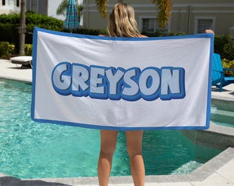 May include: A white beach towel with a blue border and the name "GREyson" in blue 3D letters.