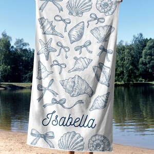 May include: White beach towel featuring blue seashell and bow illustrations. The name "Isabella" is written in a cursive font. The towel is held up against a sunny sky and a lake.