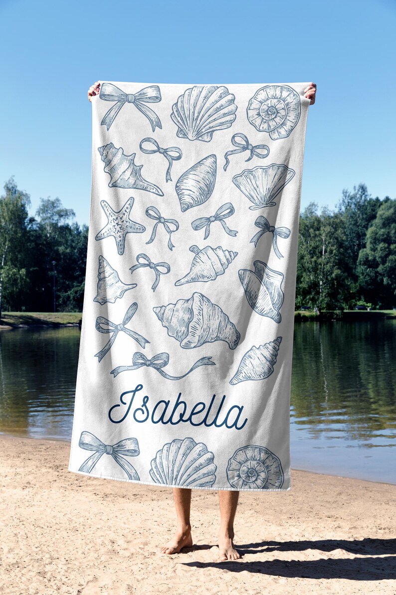May include: White beach towel featuring blue seashell and bow illustrations. The name "Isabella" is written in a cursive font. The towel is held up against a sunny sky and a lake.