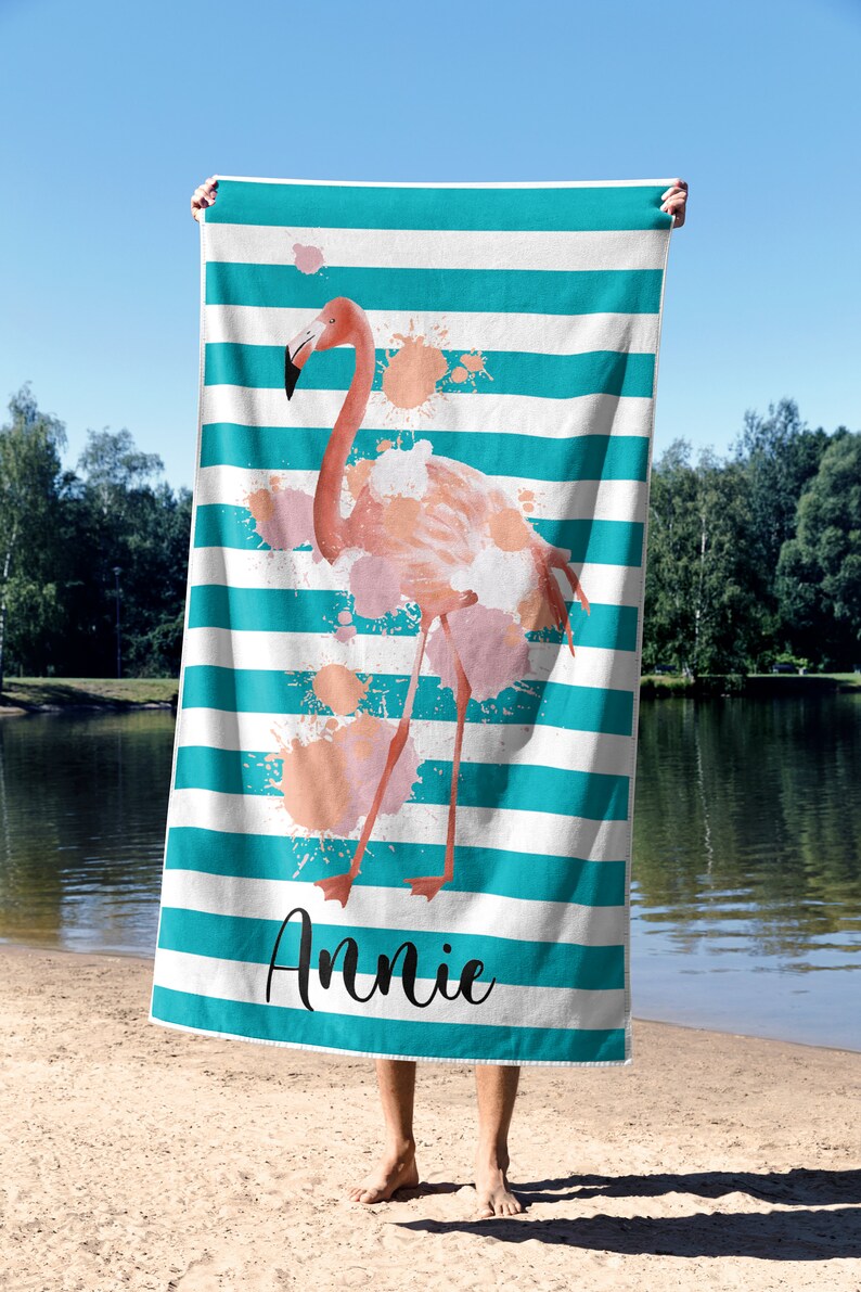 May include: A turquoise and white striped beach towel with a pink flamingo illustration and pink watercolor splatters. The towel has the name "Annie" printed on the bottom.