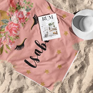 May include: Pink beach towel with a unicorn design, gold horn, floral accents, and the name "Isabella" in black script. Includes gold star accents and a white sun hat. A magazine with the word "RUM" is on the towel.