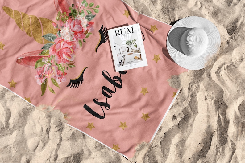 May include: Pink beach towel with a unicorn design, gold horn, floral accents, and the name "Isabella" in black script. Includes gold star accents and a white sun hat. A magazine with the word "RUM" is on the towel.