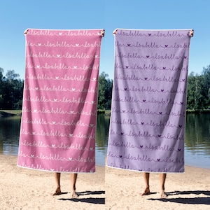 May include: Two beach towels, one pink and one purple, are held up against a blue sky and water background. Each towel is printed with the name "Isabella" in a repeating pattern with heart accents. The towels are rectangular.
