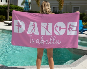 Könnte beinhalten: Ein rosa Strandtuch mit dem Wort "DANCE" in weißen Blockbuchstaben, wobei jeder Buchstabe eine Silhouette einer Ballerina zeigt. Der Name "Isabella" ist darunter in Kursivschrift geschrieben.