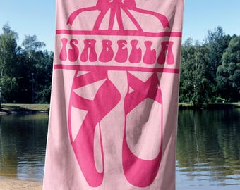Könnte beinhalten: Rosa Strandtuch mit einem Ballettschuh-Design und dem Namen "ISABELLA" in Pink. Das Handtuch hat eine Schleife oben. Das Handtuch wird an einem Strand gehalten.