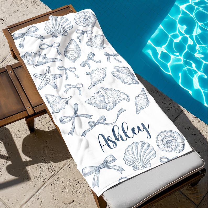 May include: White beach towel with a blue seashell and bow pattern. The name "Ashley" is printed in blue script. The towel is on a wooden lounge chair next to a pool.
