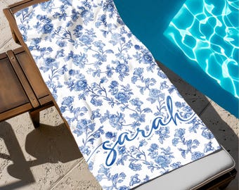Puede incluir: Toalla de playa blanca con un estampado floral azul y el nombre "Sarah" en escritura cursiva. La toalla está sobre una tumbona de madera junto a una piscina.