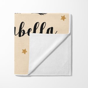 May include: A beige towel with the name "Isabella" in black script, accented with gold glitter stars. The towel is folded, revealing a white interior. A decorative item.