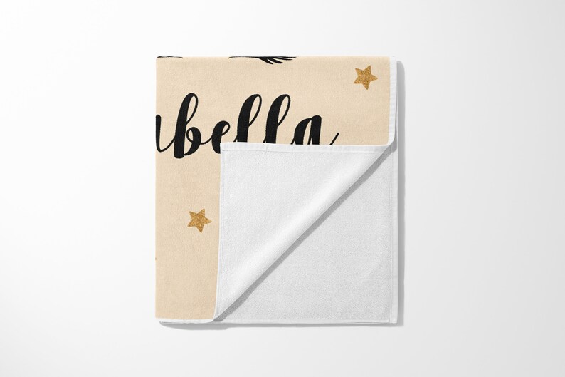 May include: A beige towel with the name "Isabella" in black script, accented with gold glitter stars. The towel is folded, revealing a white interior. A decorative item.