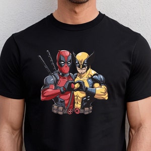 Könnte beinhalten: Schwarzes T-Shirt mit einem Aufdruck von Deadpool und Wolverine, die mit ihren Händen ein Herz bilden. Der Aufdruck ist farbig und der Text "Deadpool & Wolverine" ist unter dem Aufdruck gedruckt.