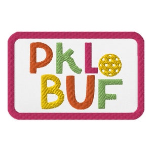 May include: A pink rectangular patch with a white background and the text "PKLO BUF" in colorful embroidered letters. A yellow embroidered pickleball is next to the text.