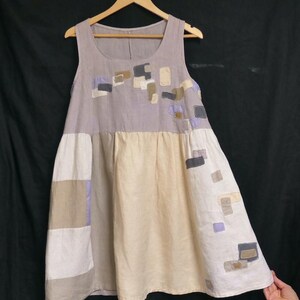 Peut inclure: Robe sans manches, longueur genou, dans des tons neutres, suspendue à un cintre en bois. La robe présente un motif patchwork avec des carrés de différentes couleurs, dont beige, gris et lavande. La robe est faite d'un tissu doux et fluide.