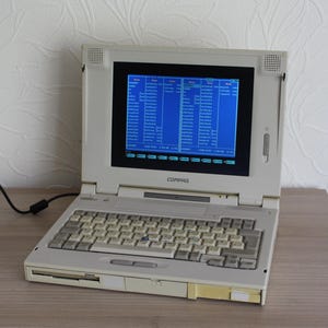 May include: An off-white Compaq laptop computer with a blue screen displaying text. The laptop has a keyboard, a floppy disk drive, and a black power cord. The screen is black-framed and the laptop is on a wooden surface.