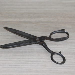 May include: A pair of vintage metal scissors with a dark, aged appearance. The blades are open, revealing their sharp edges. The handles feature finger loops for easy use. The scissors are likely used for cutting fabric or paper.