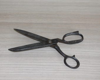 May include: A pair of vintage metal scissors with a dark, aged appearance. The blades are open, revealing their sharp edges. The handles feature finger loops for easy use. The scissors are likely used for cutting fabric or paper.