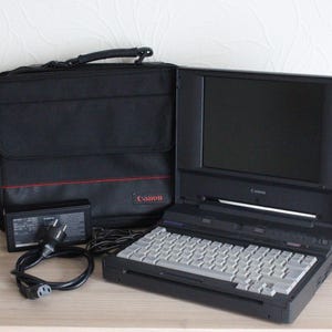 May include: A vintage Canon laptop computer, black carrying case, power adapter, and cable. The laptop has a keyboard and a screen. The carrying case is black with a red stripe and the Canon logo. The power adapter and cable are black.