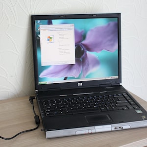 Op de afbeelding: Zwarte HP laptop met zilveren rand, open op een houten oppervlak. Het scherm toont een Windows-interface met een paarse bloemenachtergrond. Een zwart netsnoer is aangesloten op de laptop.