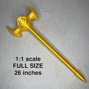 May include: A full-size, gold-coloured prop weapon, measuring 66 cm in length. The prop features a central eye-shaped design with curved blades on either side of the head. The text "1:1 scale FULL SIZE 26 inches" is visible.