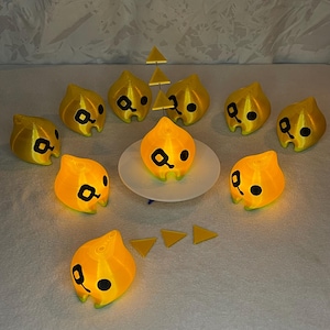 Puede incluir: Un grupo de nueve figuras amarillas impresas en 3D con ojos negros y un solo ojo negro a cada lado de su boca. Las figuras están dispuestas en círculo alrededor de un plato blanco con una sola figura en el plato. Hay tres formas triangulares amarillas en el fondo.