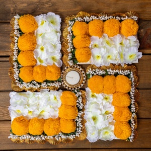 Puede incluir: Un diseño tradicional de rangoli indio hecho con flores de caléndula y flores blancas, dispuestas en forma de cuadrado con una pequeña vela en el centro.