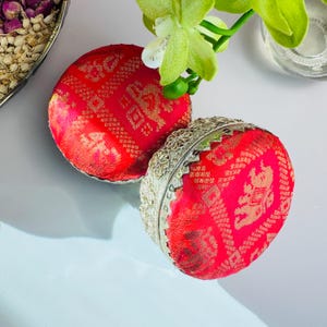 May include: Two red and gold decorative boxes with intricate patterns. The boxes are round and have a silver trim.