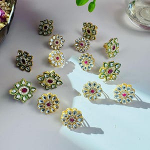 May include: A collection of 14 gold-tone rings with green and white enamel and red gemstones. The rings are arranged on a white surface.