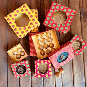 May include: An overhead shot of colorful gift boxes with a floral pattern. Some boxes are open, revealing treats inside. The boxes are yellow, red, and pink, with a cutout of a deity. The treats are round and wrapped in gold foil.