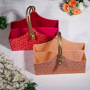 May include: Two decorative gift baskets with gold handles. The basket on the left is pink with gold floral patterns. The basket on the right is peach with gold floral patterns.
