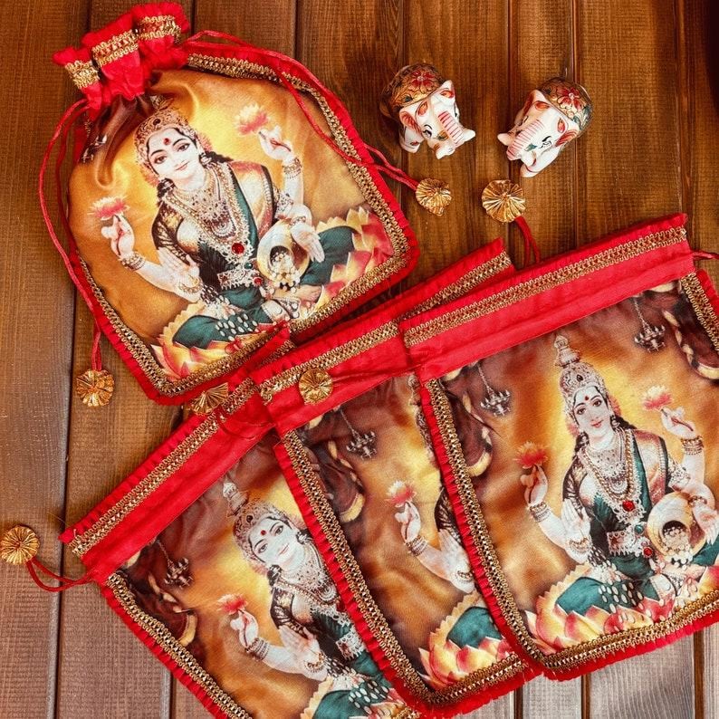 K&ouml;nnte beinhalten: Vier rote Kordelbeutel mit goldenen Verzierungen und einem gedruckten Bild einer hinduistischen G&ouml;ttin. Die Beutel sind pyramidenf&ouml;rmig auf einer Holzoberfl&auml;che angeordnet.