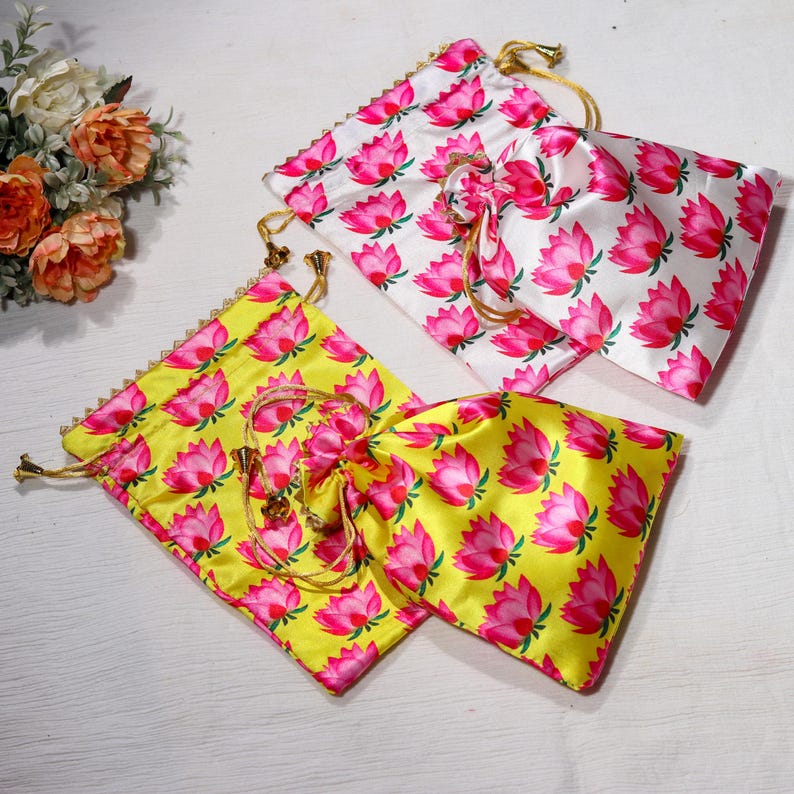 K&ouml;nnte beinhalten: Zwei Kordelbeutel mit rosa Lotusblumenmustern. Ein Beutel ist wei&szlig; mit rosa Blumen und der andere ist gelb mit rosa Blumen.