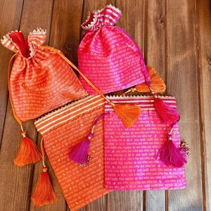 K&ouml;nnte beinhalten: Vier kleine Kordelbeutel mit Quasten. Die Beutel sind aus rosa und orangefarbenem Stoff mit goldenen Verzierungen und mit gedrucktem Text verziert.