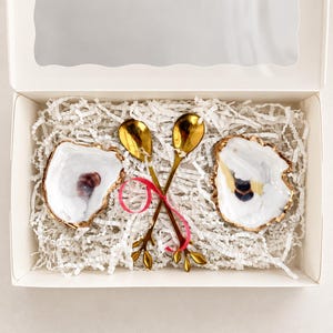 May include: A white gift box containing two white oyster shell dishes with gold-painted edges and two gold-toned metal spoons with branch-shaped handles, tied together with a thin pink ribbon and nestled in white shredded paper filler.