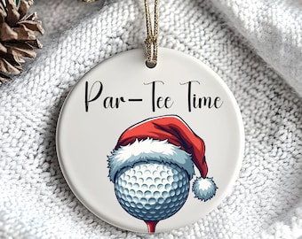 Puede incluir: Adorno navideño redondo y blanco con la frase "Par-Tee Time". El adorno muestra una pelota de golf con un gorro de Papá Noel. Cuelga de un cordón dorado. El fondo incluye piñas y ramas de abeto.