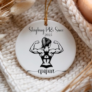 May include: White ceramic ornament with a gold string for hanging. The ornament features a black graphic of a muscular person flexing, with the text "Sleighing PRs Since 2025" and the name "emma".