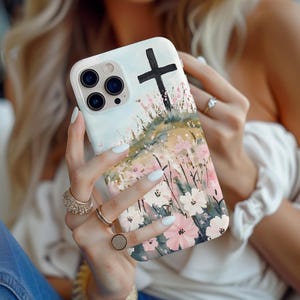 Puede incluir: Una funda blanca para iPhone con un diseño de acuarela de flores rosas y una cruz negra. La funda tiene un recorte para el objetivo de la cámara.