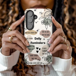 May include: A white phone case with a floral and geometric design. The case features text including "Daily Reminders", "Bad Bitch Energy Only", and "Take No Shit, Give No Fucks". The case has a camera module with three lenses. The case is held by a person.