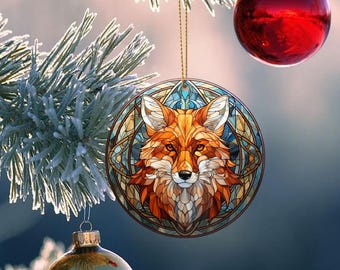 May include: A round Christmas ornament featuring a detailed fox design. The fox has orange and white fur, set against a stained-glass style background with blue and gold accents. The ornament hangs from a gold string, with a red ball ornament and frosted pine branches in the background.