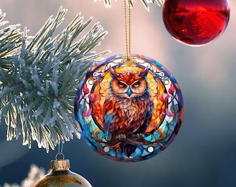 May include: A stained glass ornament featuring a colorful owl with a detailed design. The owl is perched on a branch with flowers and leaves surrounding it. The ornament is round and has a gold hanger.