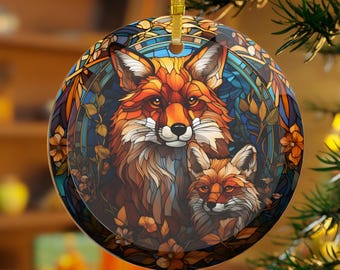 May include: Round ceramic ornament featuring a detailed illustration of two foxes. The artwork uses warm colors like orange, brown, and gold, with a stained-glass effect. The ornament has a gold ribbon for hanging and is surrounded by floral elements.