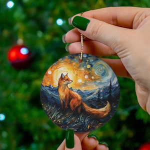 Puede incluir: Adorno navideño redondo con un zorro en un paisaje con un cielo estrellado y un sol brillante. El zorro es naranja y blanco, y el fondo tiene tonos de azul, naranja y amarillo. El adorno se sujeta con una cuerda dorada.