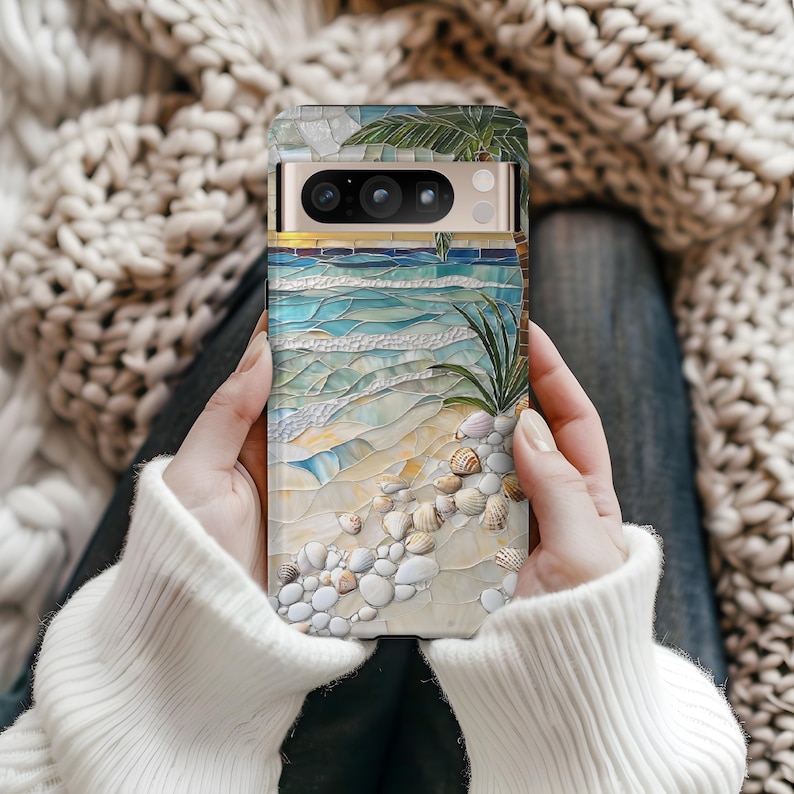 May include: A phone case featuring a beach scene mosaic design. The case showcases a sandy shore, ocean waves, seashells, and palm trees in shades of blue, beige, and white. The case is held in a person's hands.